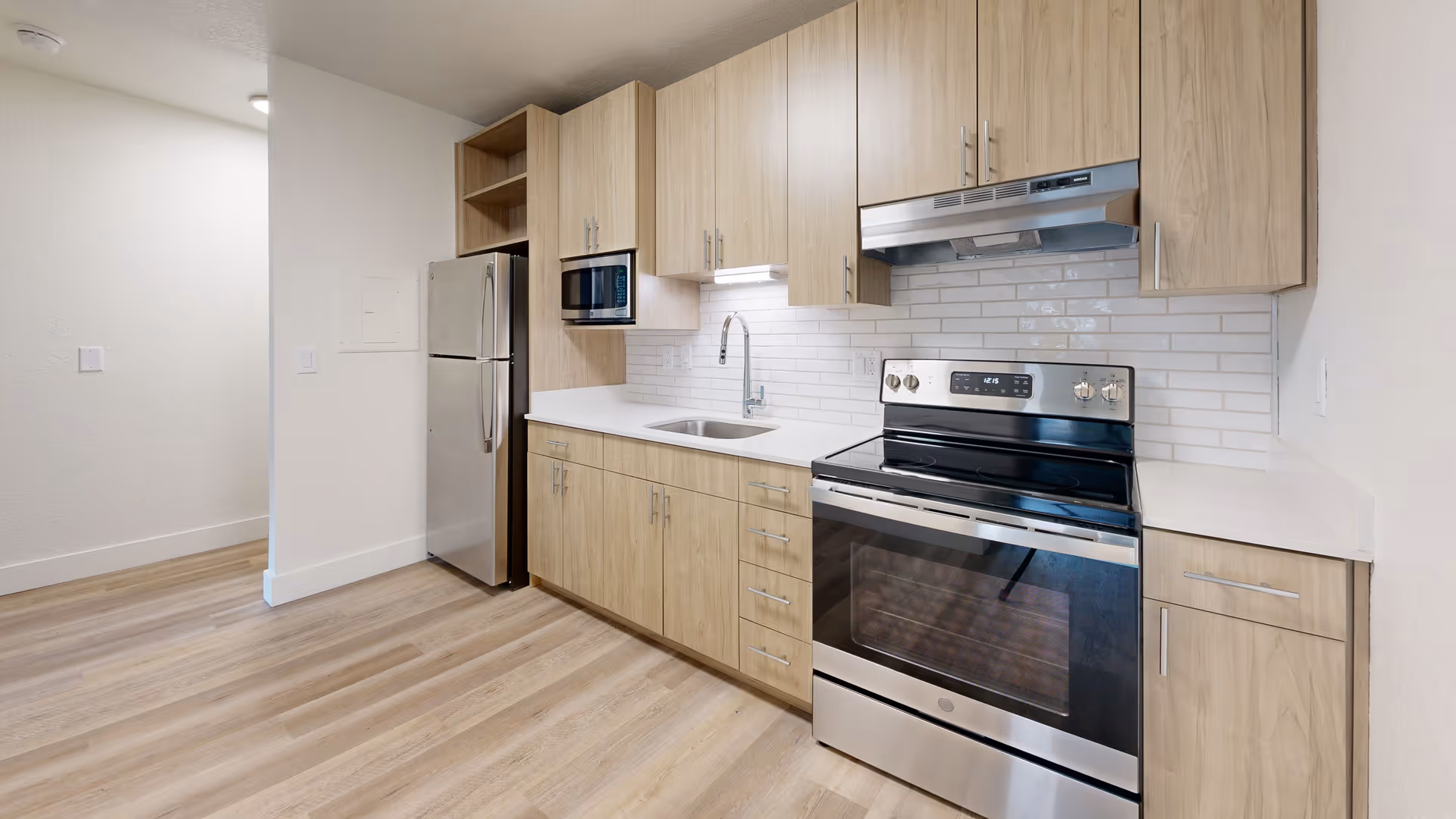 Modern kitchen with light wood cabinets, stainless steel refrigerator, stove and microwave, a sink, and white subway tile backsplash.