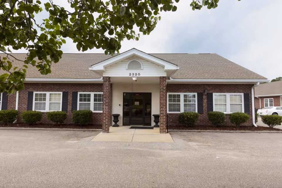 Front exterior view of a single-story brick building with white trim and black shutters. The building has a covered entrance with two brick columns and the number 2320 above the door. There are bushes along the front and a tree branch partially visible at the top left corner.