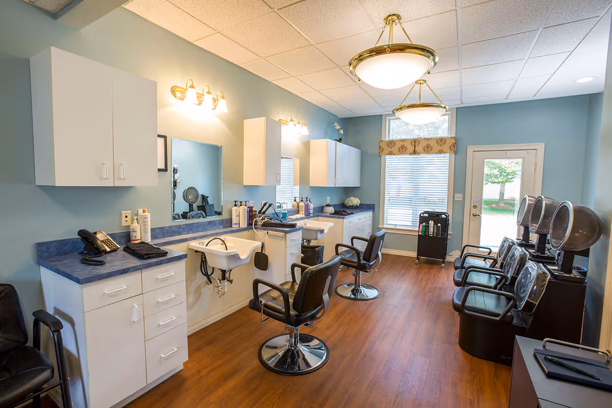 Interior of a senior living facility hair salon with two styling chairs in front of sinks, mirrors, and cabinets. There are three black hair drying chairs with hooded dryers along the right wall, wooden flooring, and a window with blinds and a valance letting in natural light.