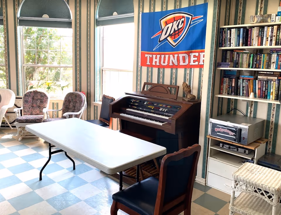 Sunlit common room with a folding table and chairs, an electronic organ, bookshelf and an OKC Thunder flag on the wall.