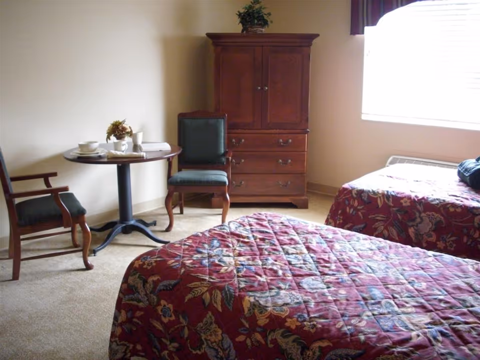A bedroom with two beds covered in red floral patterned bedspreads. There is a wooden cabinet with drawers against the wall and a small round table with two chairs. The room has a window with blinds partially open, allowing natural light to enter.