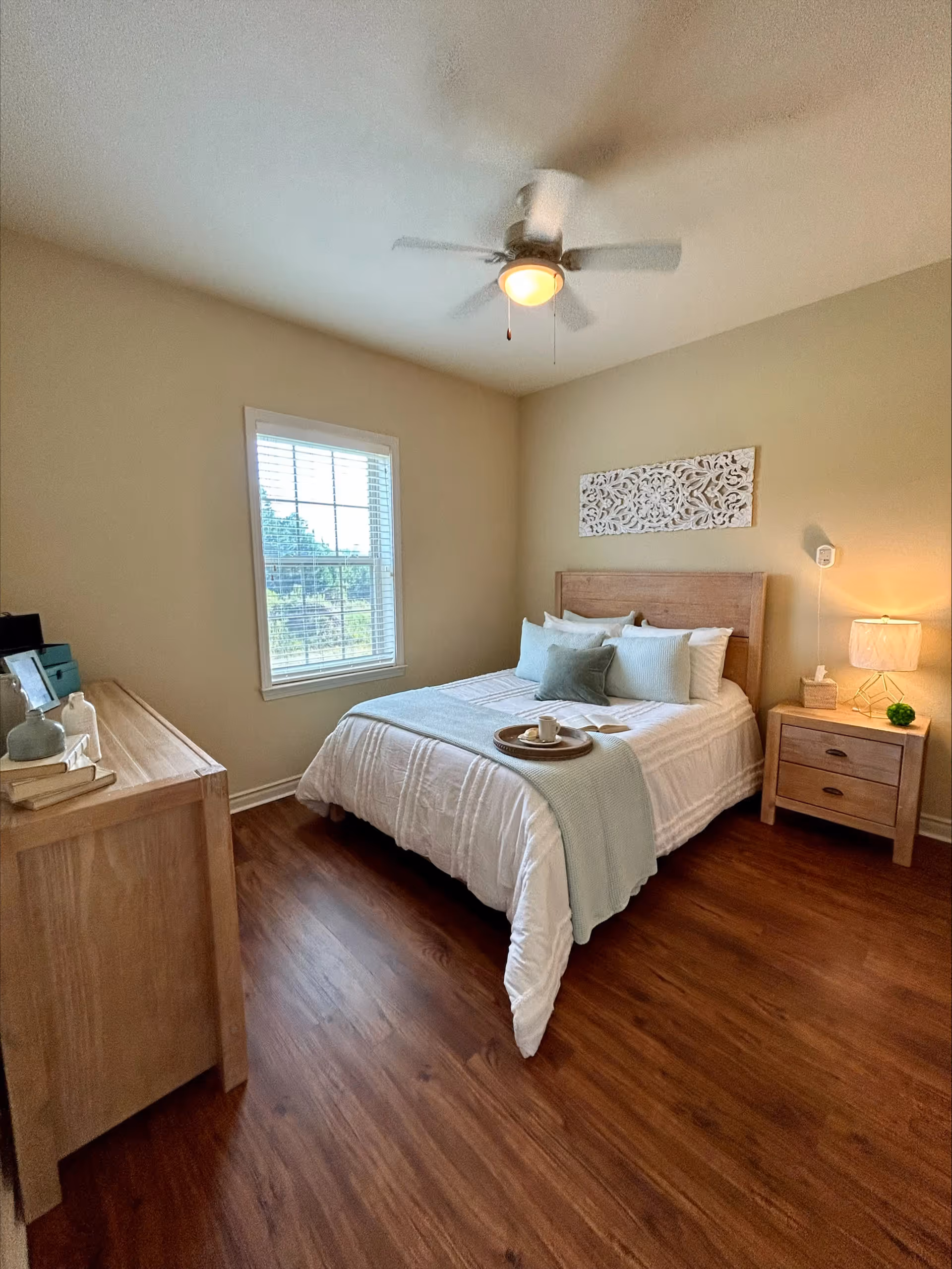 A cozy bedroom with a wooden bed frame and white bedding accented with light blue pillows and a throw blanket. A wooden nightstand with a lamp and decorative items is beside the bed. There is a wooden dresser with books and decor on the left side of the room. A window with white blinds lets in natural light, and a ceiling fan with a light is mounted on the ceiling. The floor is a warm wood tone.