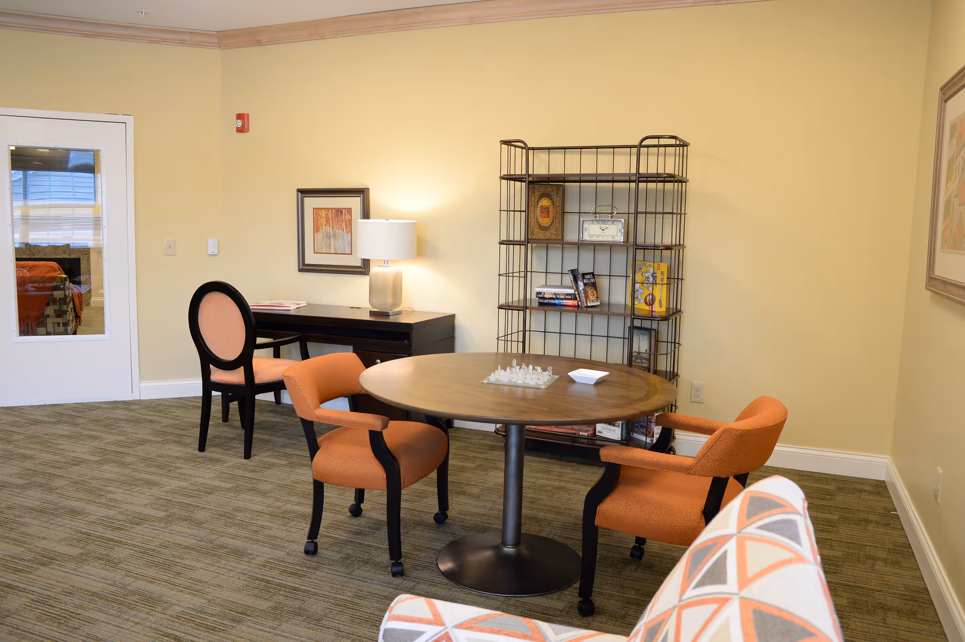 A cozy room with a round wooden table surrounded by two orange armchairs. A chess set is placed on the table. In the background, there is a black desk with a lamp and a chair with a peach-colored cushion. A metal bookshelf with books and decorative items is against the wall. The walls are painted light yellow, and the floor is carpeted in a neutral tone. A patterned armchair is partially visible in the foreground.