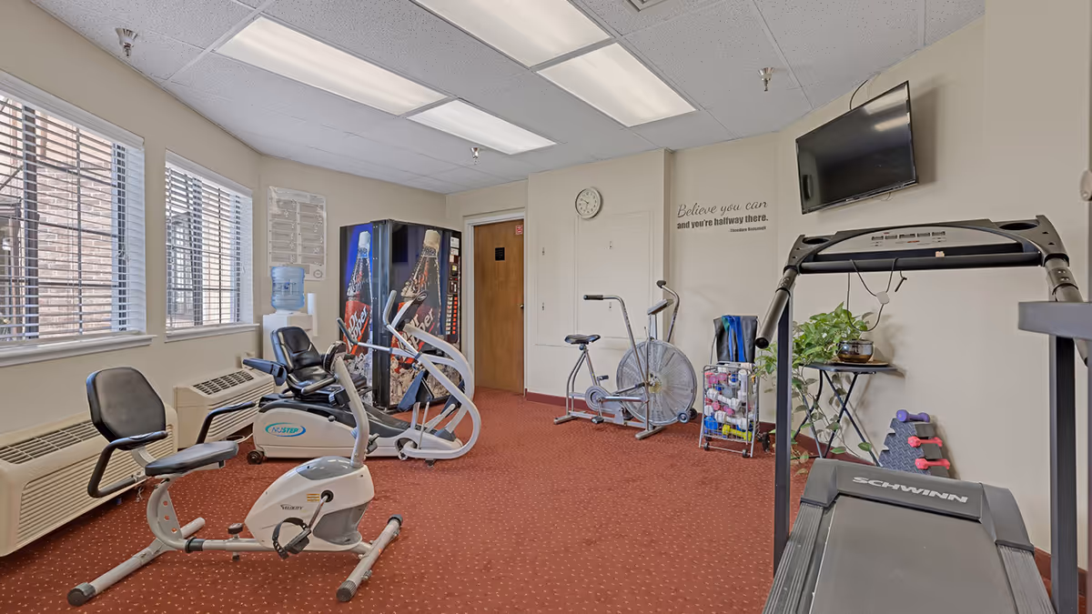 Small fitness room with a treadmill, stationary bikes, vending machine, water cooler, and wall-mounted TV.