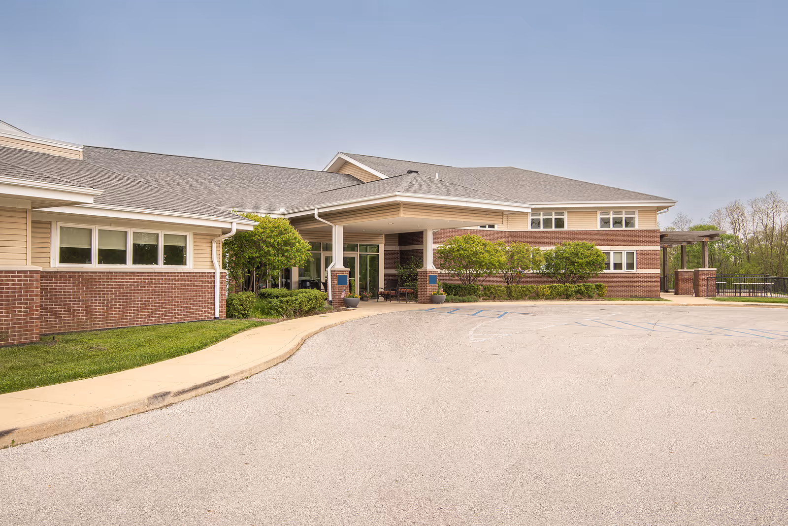 Front entrance of a single-story brick-and-siding senior living facility with a covered drop-off, shrubs, and an empty paved driveway.