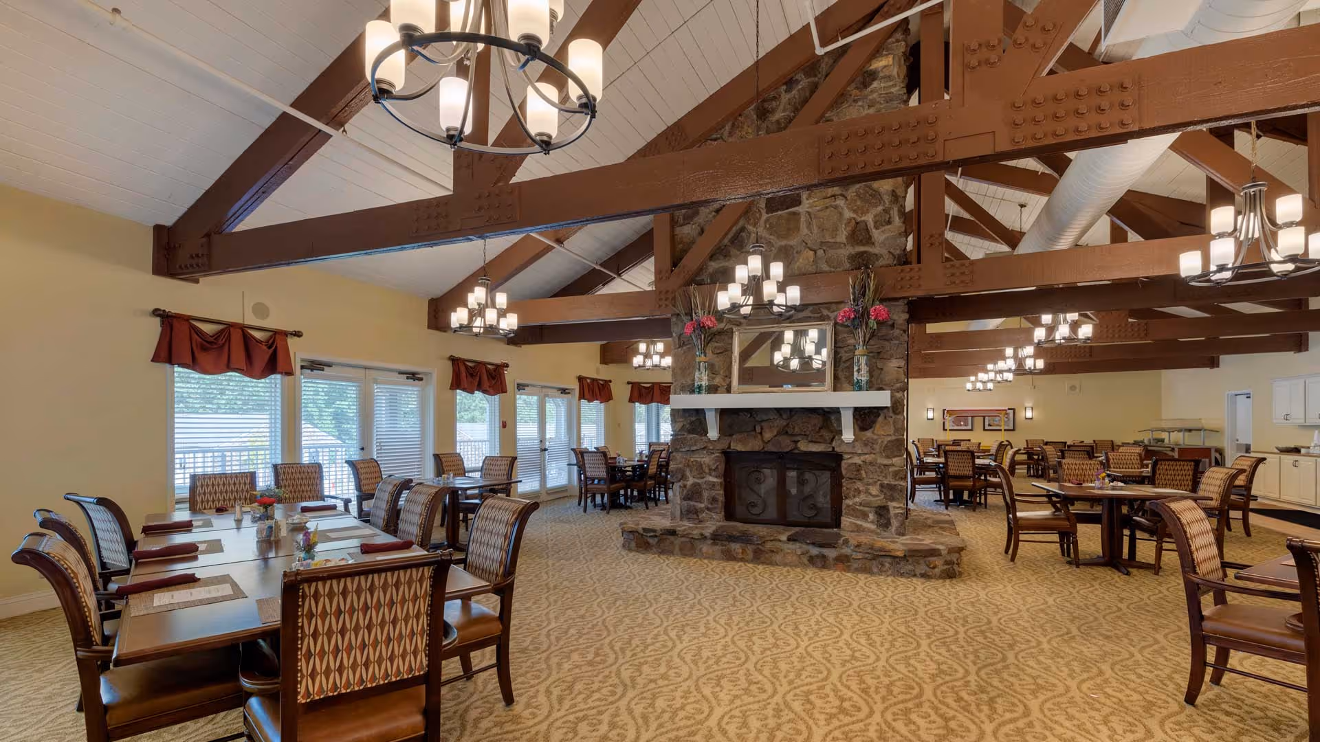 A spacious dining room with multiple wooden tables and cushioned chairs arranged neatly. The room features a large stone fireplace in the center with a mirror and floral decorations on the mantel. Exposed wooden beams and multiple chandeliers hang from the ceiling, and large windows with red valances allow natural light to fill the space.