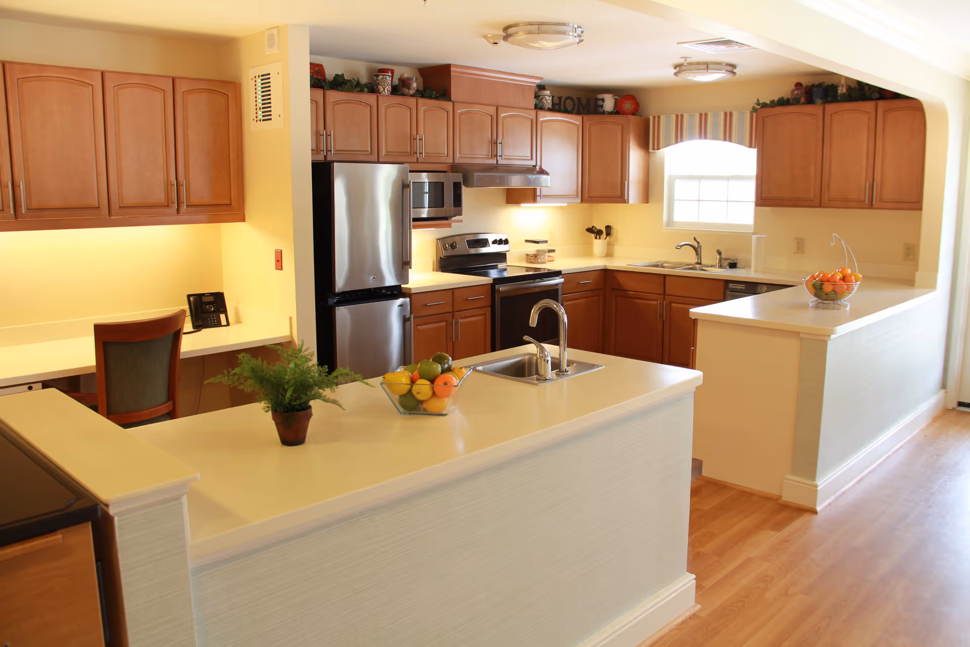 A bright, spacious kitchen with wooden cabinets, stainless steel refrigerator, stove, and microwave. The kitchen features two white countertops with sinks, a bowl of fruit, and a small potted plant. There is a window with a striped valance letting in natural light and a small desk area with a chair and phone on the left side.