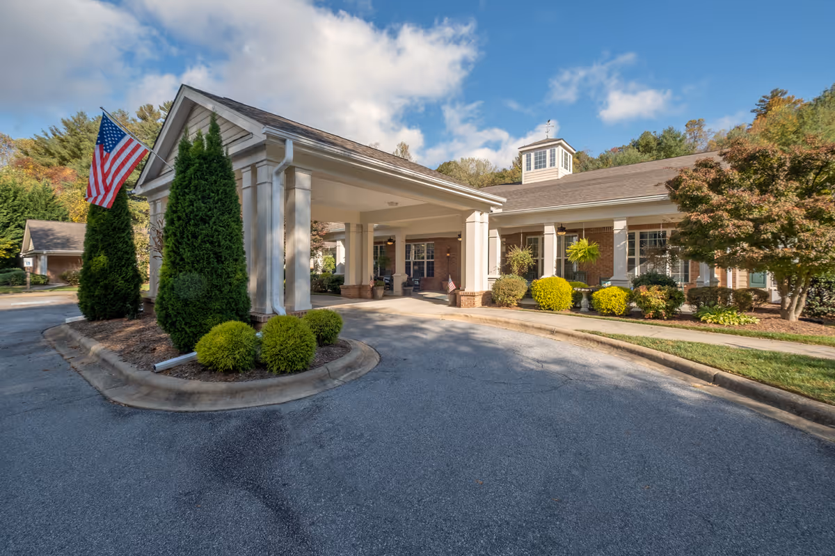 Entrance porte-cochere of a brick senior living building with an American flag, shrubs, and a blue sky.