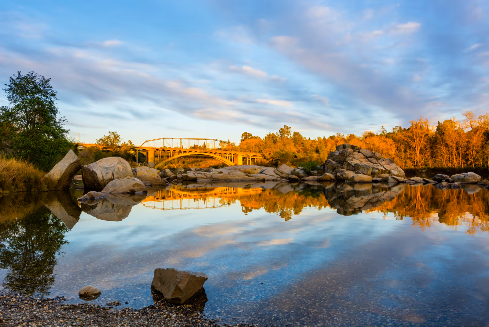 A serene outdoor scene featuring a calm river with large rocks and trees on both sides. A bridge spans the river in the background, and the sky is partly cloudy with soft sunlight illuminating the landscape.