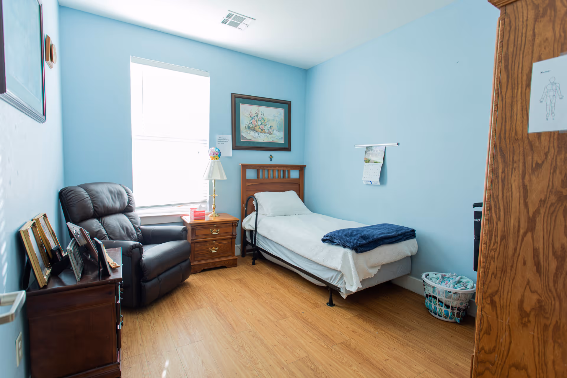 A small bedroom with light blue walls and wooden flooring. The room contains a single bed with white bedding and a folded blue blanket on top, a wooden nightstand with a lamp and some items, a black recliner chair, a wooden dresser with framed photos, and a laundry basket on the floor. There is a window letting in natural light, a framed picture on the wall above the bed, and a calendar hanging on the wall.