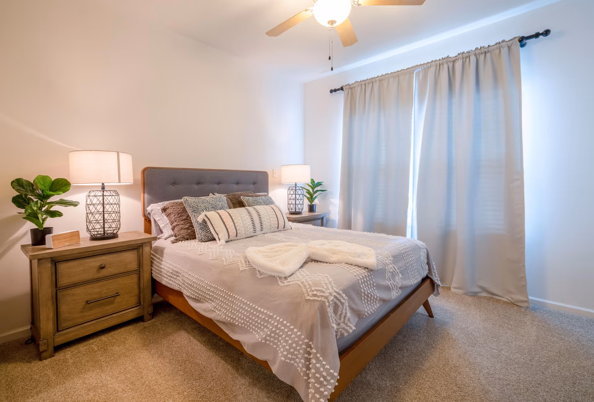 Well-lit bedroom with a neatly made bed, two nightstands with lamps and plants, a ceiling fan, and closed curtains.