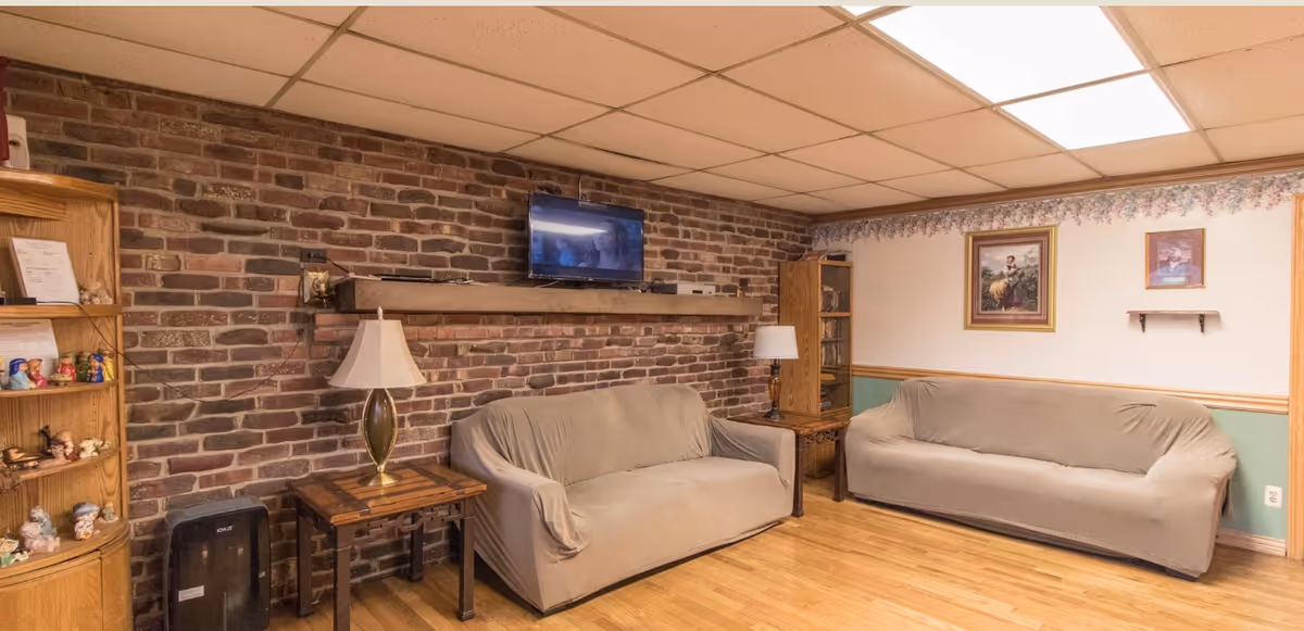 A cozy living room with two beige slipcovered sofas facing each other, a brick accent wall with a mounted flat-screen TV, wooden side tables with lamps, a wooden bookshelf with decorative items, and framed artwork on the opposite wall. The room has a wood floor and a drop ceiling with fluorescent lighting.