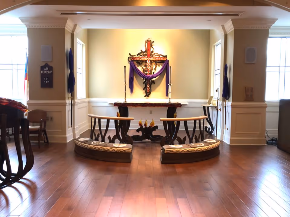 Interior view of a chapel or prayer room with wooden flooring, a decorative cross draped with purple cloth on the wall, two tall candlesticks on a wooden altar, and curved wooden kneelers with cushions in front of the altar. The room has white wainscoting and two windows letting in natural light.