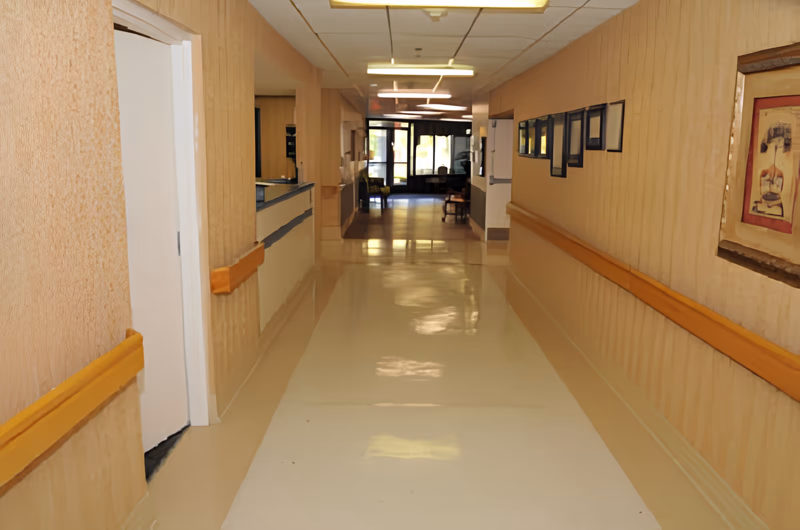A clean, well-lit hallway in a senior living facility with beige walls, wooden handrails on both sides, framed pictures on the right wall, and a reception window on the left. At the end of the hallway, there is a glass door entrance with chairs visible near the door.
