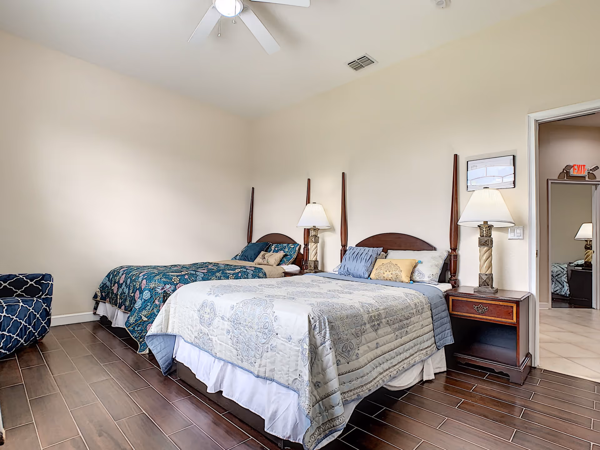 A bedroom with two beds, one with a blue and white patterned bedspread and the other with a dark floral bedspread. There are two bedside tables with lamps, a blue patterned armchair, and a ceiling fan. The room has wooden flooring and beige walls. An open door reveals a hallway with a visible exit sign.
