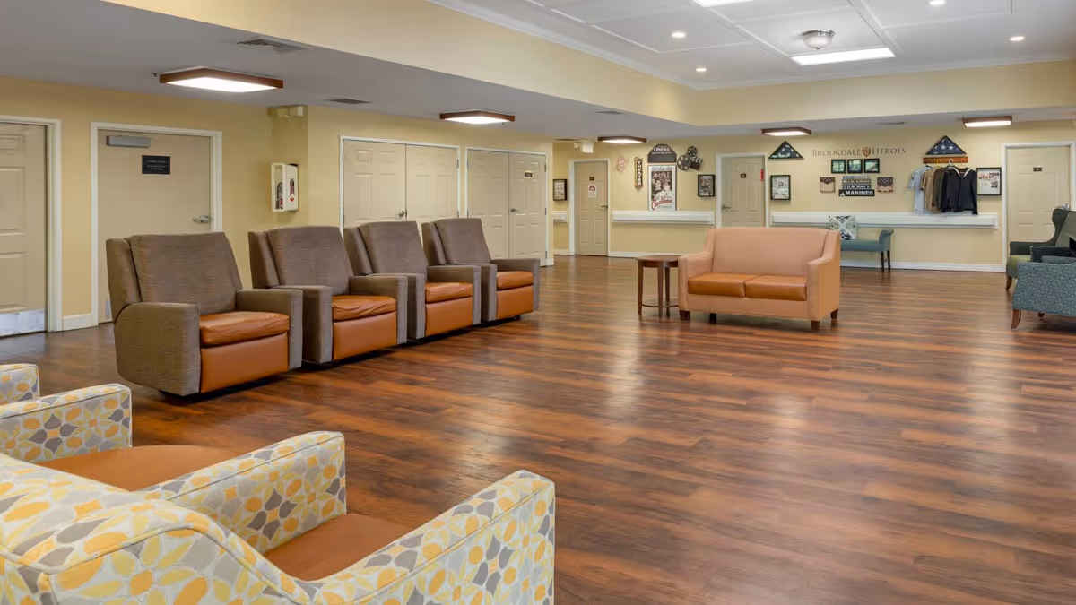 A spacious senior living facility common area with multiple armchairs and sofas arranged on a wood floor. The walls are light yellow with several doors and a reception desk in the background. Various decorations and plaques are displayed on the wall behind the desk.