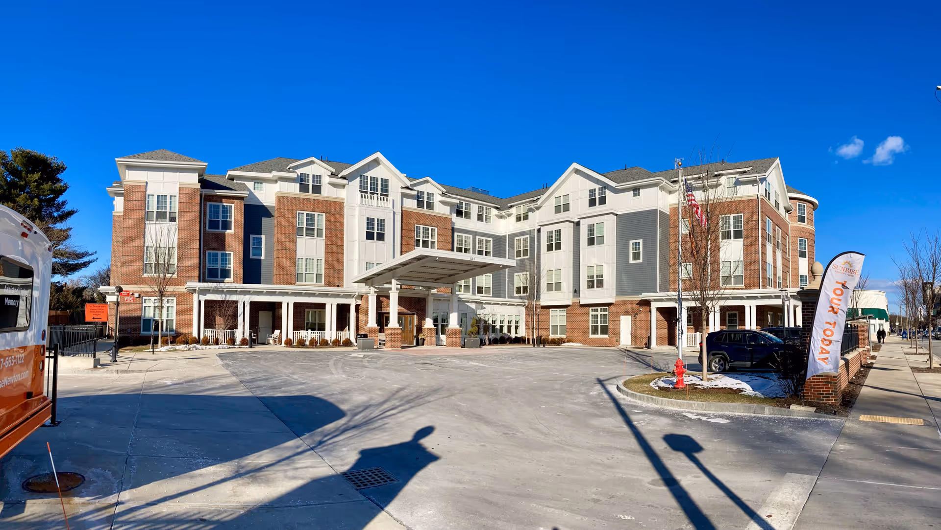 Exterior view of a multi-story senior living facility building with a covered entrance, brick and white siding facade, an American flag, a 'Tour Today' flag, and a clear blue sky.