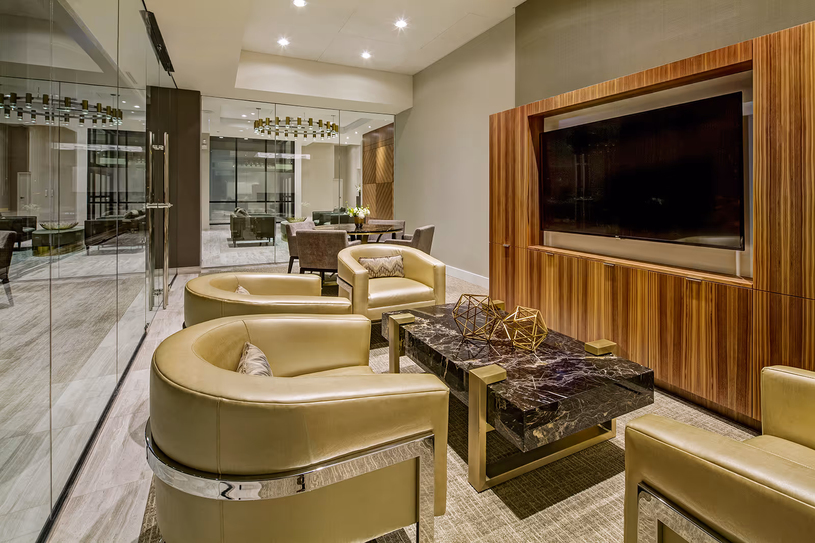 Modern lounge with curved leather armchairs, a marble coffee table and large wall-mounted TV in a glass-walled common area.