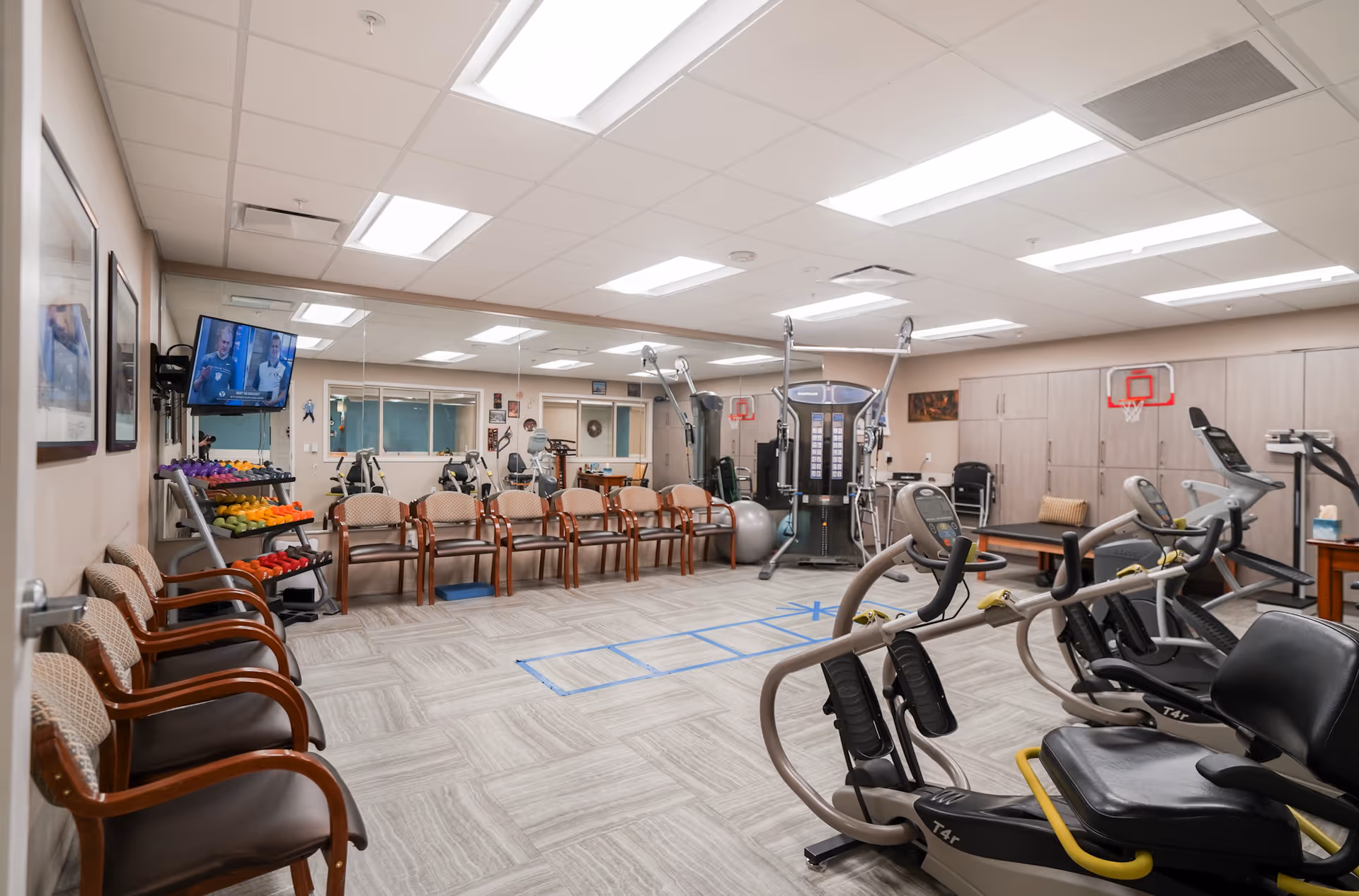 Well-lit senior exercise room with cardio and strength machines, a rack of weights, chairs along the wall and a large mirror.