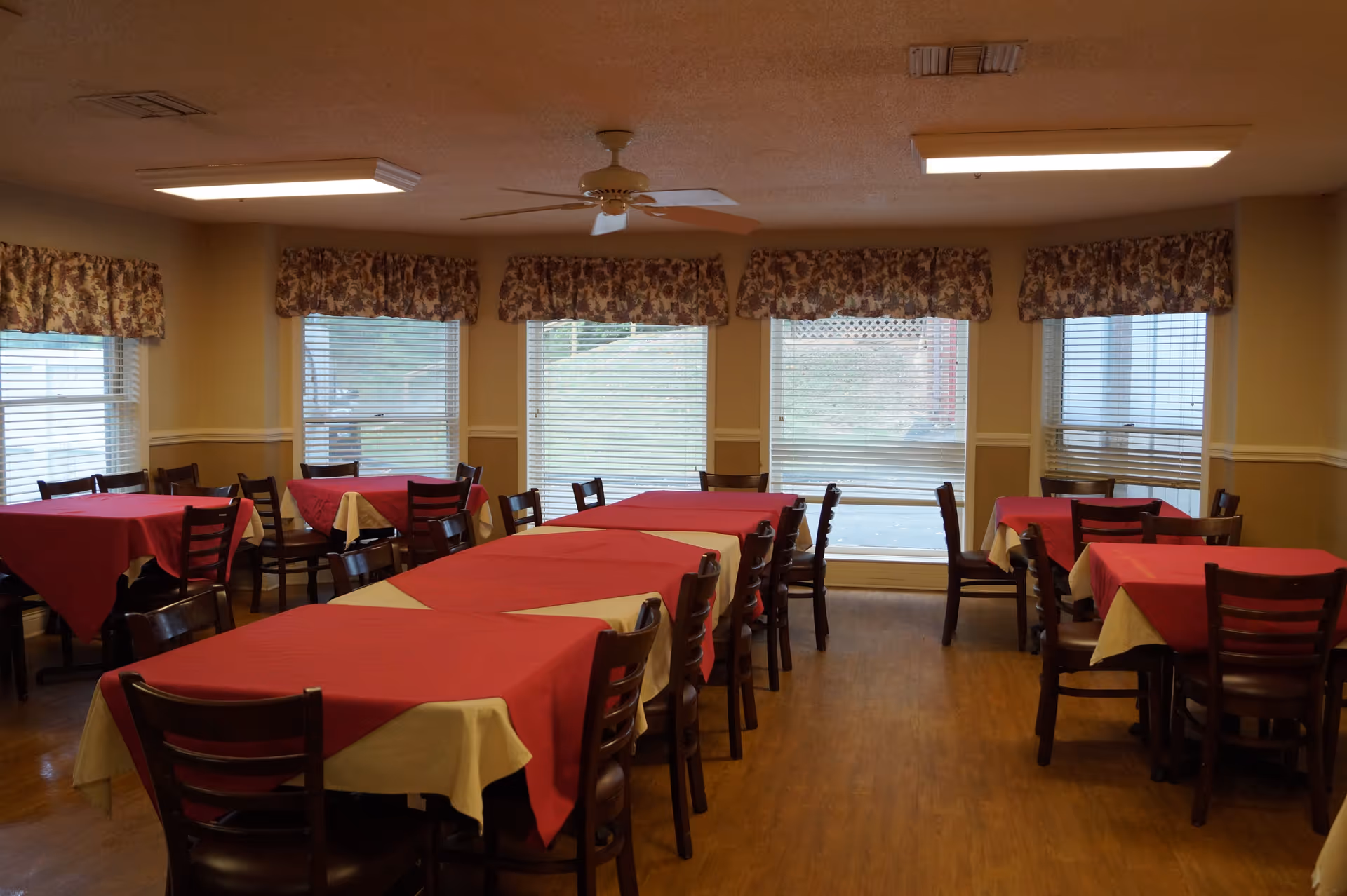 Dining room with multiple tables covered with red and beige tablecloths, surrounded by wooden chairs. The room has large windows with floral valances and blinds, a ceiling fan, and overhead fluorescent lights. The floor is wooden.
