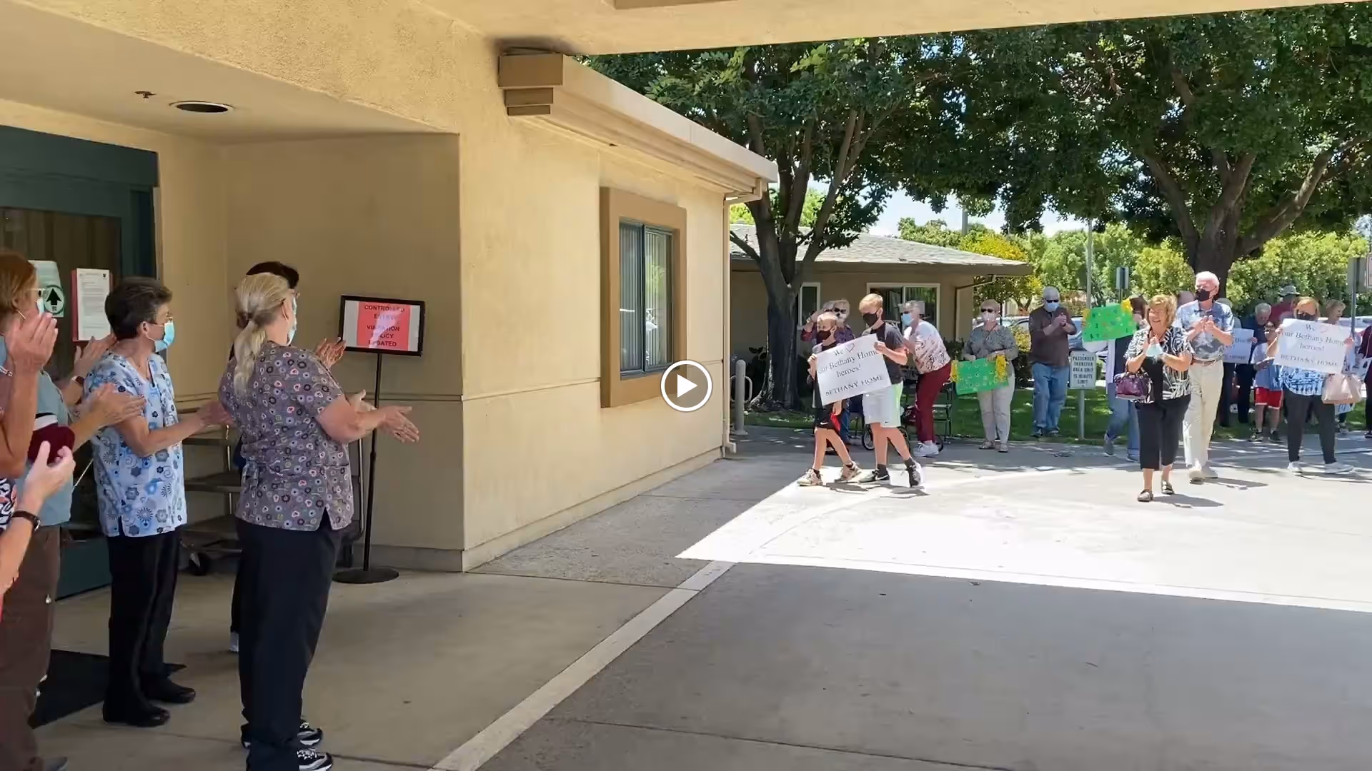A group of people standing outside a building, some wearing masks and clapping, while others hold signs and walk towards the building entrance under a shaded area with trees in the background.