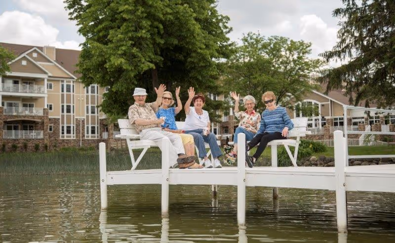 Five elderly people sitting on a white wooden dock by the water, waving and smiling. Behind them are trees and a multi-story residential building with balconies.