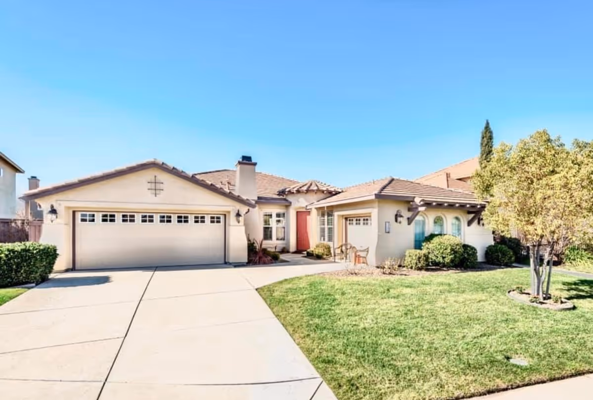 Front exterior view of a single-story house with a two-car garage, a driveway, a well-maintained lawn, bushes, and a tree under a clear blue sky.