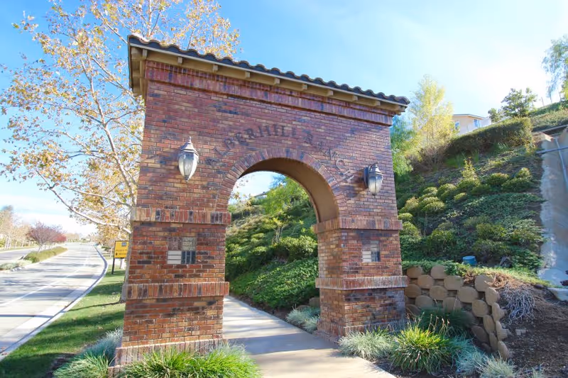 Brick arched entryway with lanterns leading to a landscaped sidewalk along a hillside under a clear sky.