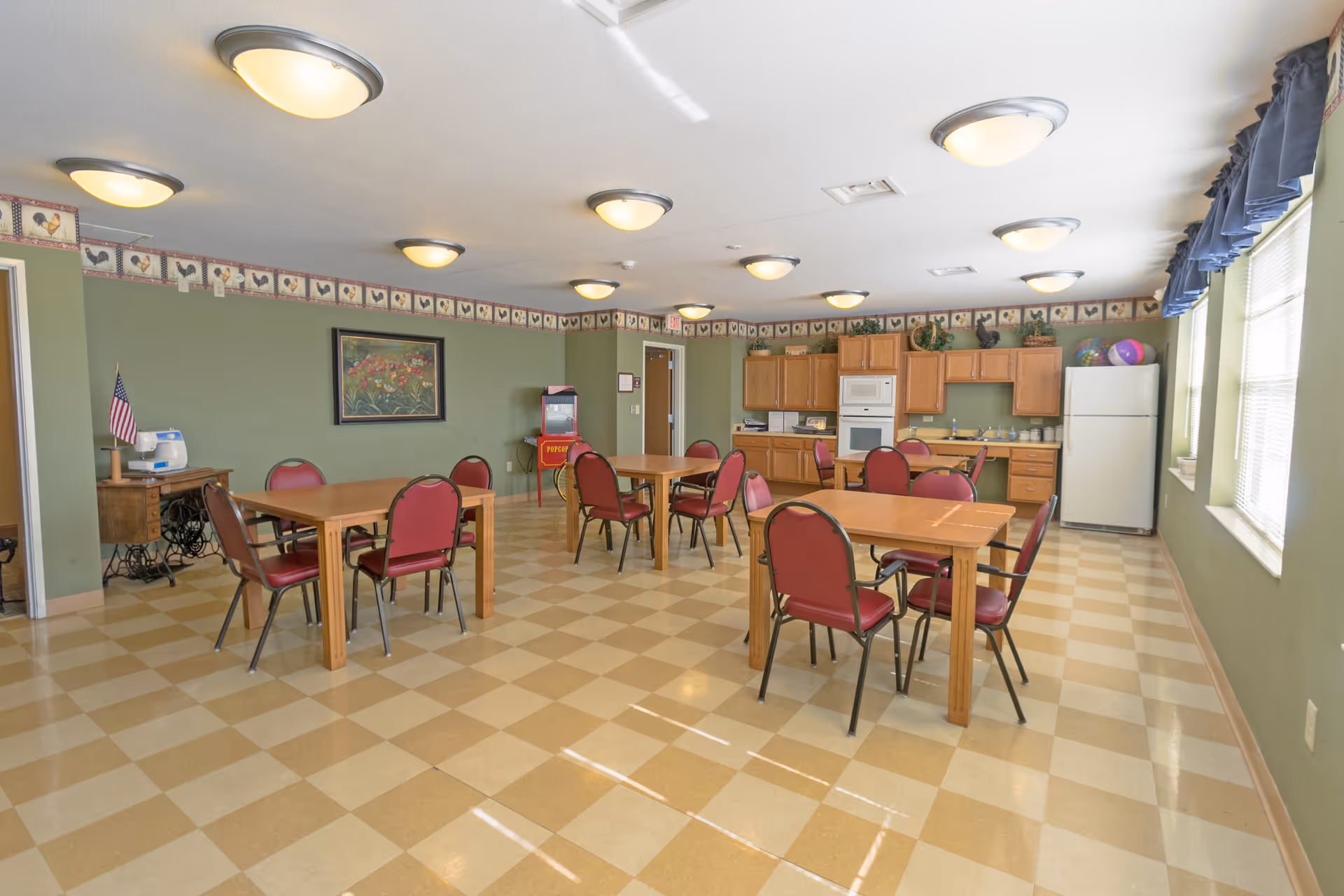 A spacious dining room with several wooden tables and red cushioned chairs arranged neatly. The room has green walls with a decorative border near the ceiling, multiple ceiling lights, and large windows with blue valances letting in natural light. At the far end, there is a kitchenette area with wooden cabinets, a white refrigerator, microwave, and sink. A popcorn machine and a small table with an American flag are also visible.