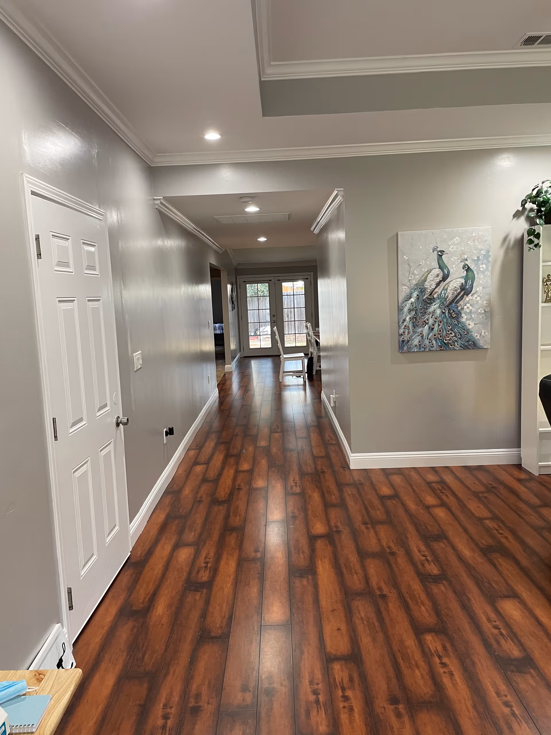 A long hallway with wooden flooring and light gray walls. On the left side, there is a closed white door and a small table with some items on it. On the right side, there is a wall with a painting of two peacocks and a white shelving unit with a plant on top. At the end of the hallway, there are glass double doors leading outside, with a glimpse of a fenced yard. The ceiling has recessed lighting and crown molding.