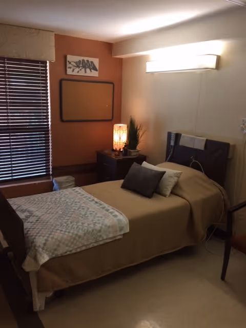 Single-bed nursing facility bedroom with a nightstand, lamp, pillows, and window blinds.