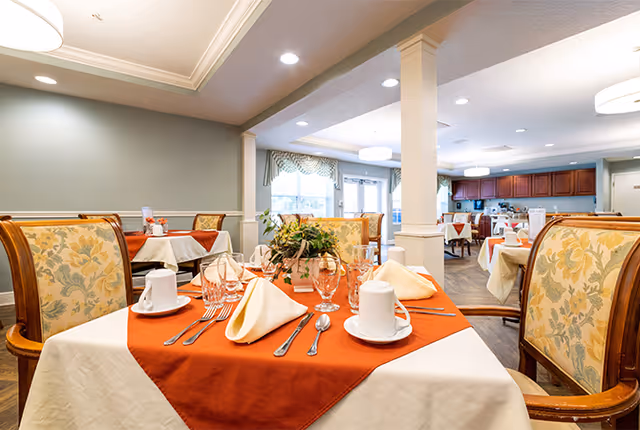 A dining room in a senior living facility with tables set for a meal. The tables have white tablecloths with orange overlays, neatly folded napkins, cups, glasses, and silverware. The chairs have floral upholstery. The room is well-lit with ceiling lights and has large windows with curtains allowing natural light.