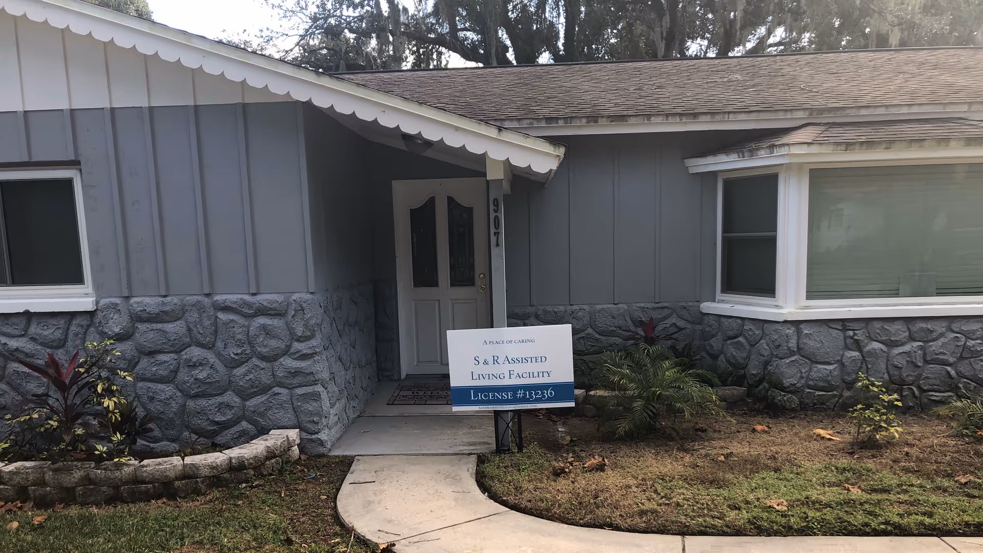 Front entrance of a single-story building with gray stone and siding exterior, a white door, and a small garden area with plants. A sign in front reads 'S & R Assisted Living Facility License #13236'.