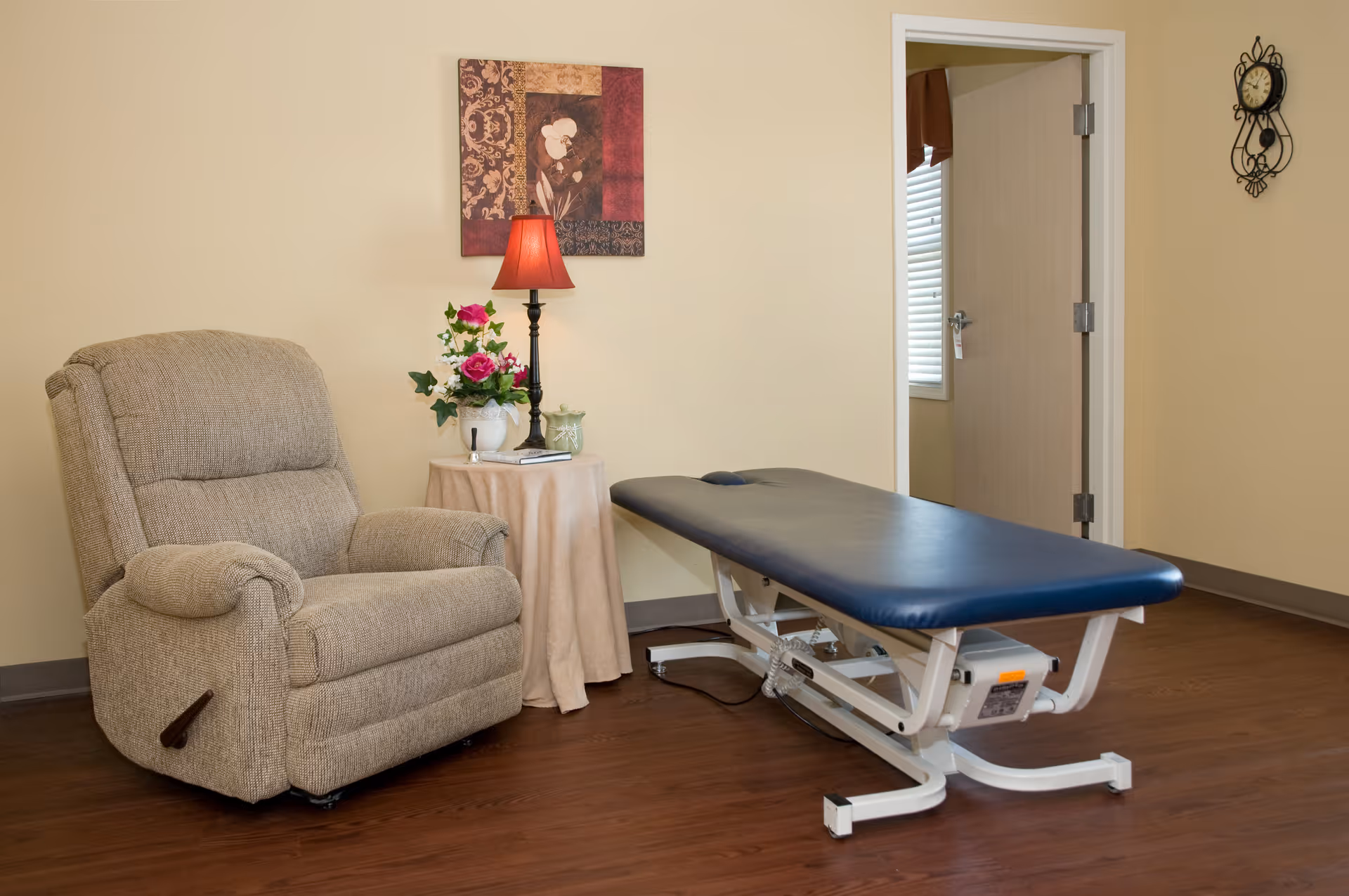 A cozy interior room with a recliner, a side table with a lamp and flowers, and a blue therapy/exam table.