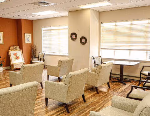 A bright and cozy common area with several beige upholstered armchairs arranged around small tables. The room has large windows with blinds allowing natural light to fill the space, wooden flooring, and warm-toned walls with framed artwork and decorative items.