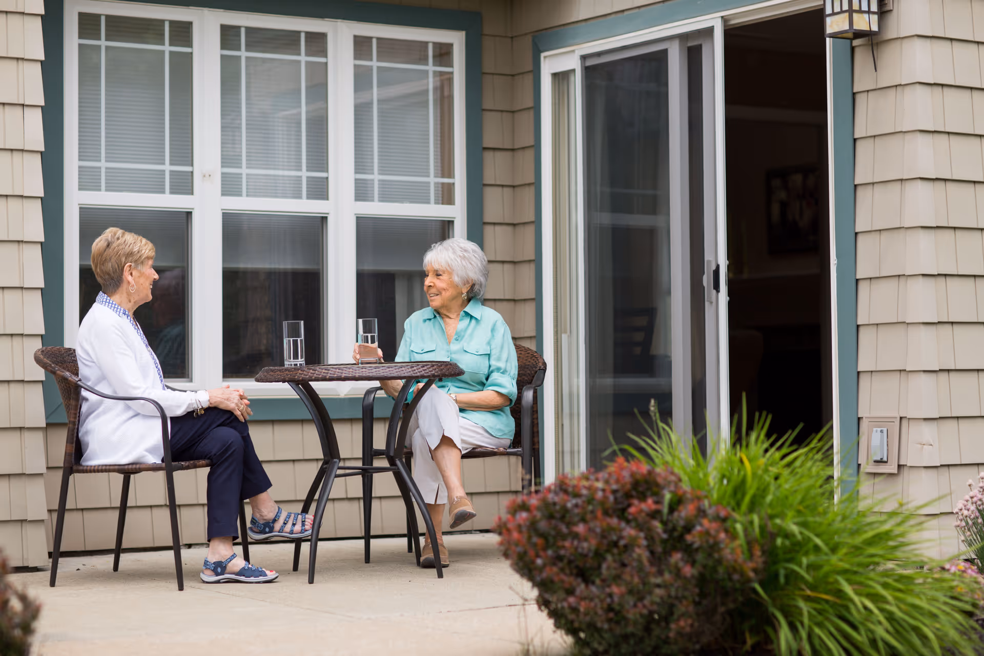 Two elderly women sitting outside on a patio at a small round table with glasses of water, engaged in conversation. The patio is adjacent to a building with beige siding, large windows, and a sliding glass door. There are green plants and bushes near the patio.