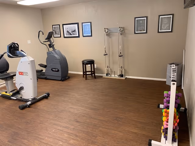 A small exercise room with wooden flooring and beige walls. The room contains exercise equipment including a recumbent bike, an elliptical machine, a resistance training apparatus, a stool, and a rack with colorful dumbbells. Several framed pictures and diagrams are hung on the walls.