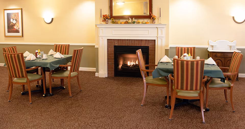 A cozy dining area with two tables covered in green tablecloths, each surrounded by four wooden chairs with striped upholstery. The room features a lit fireplace with a white mantel decorated with small pumpkins and autumn-themed items. There are wall sconces providing warm lighting, a framed floral painting on the left wall, and a wooden sideboard on the right side of the room.