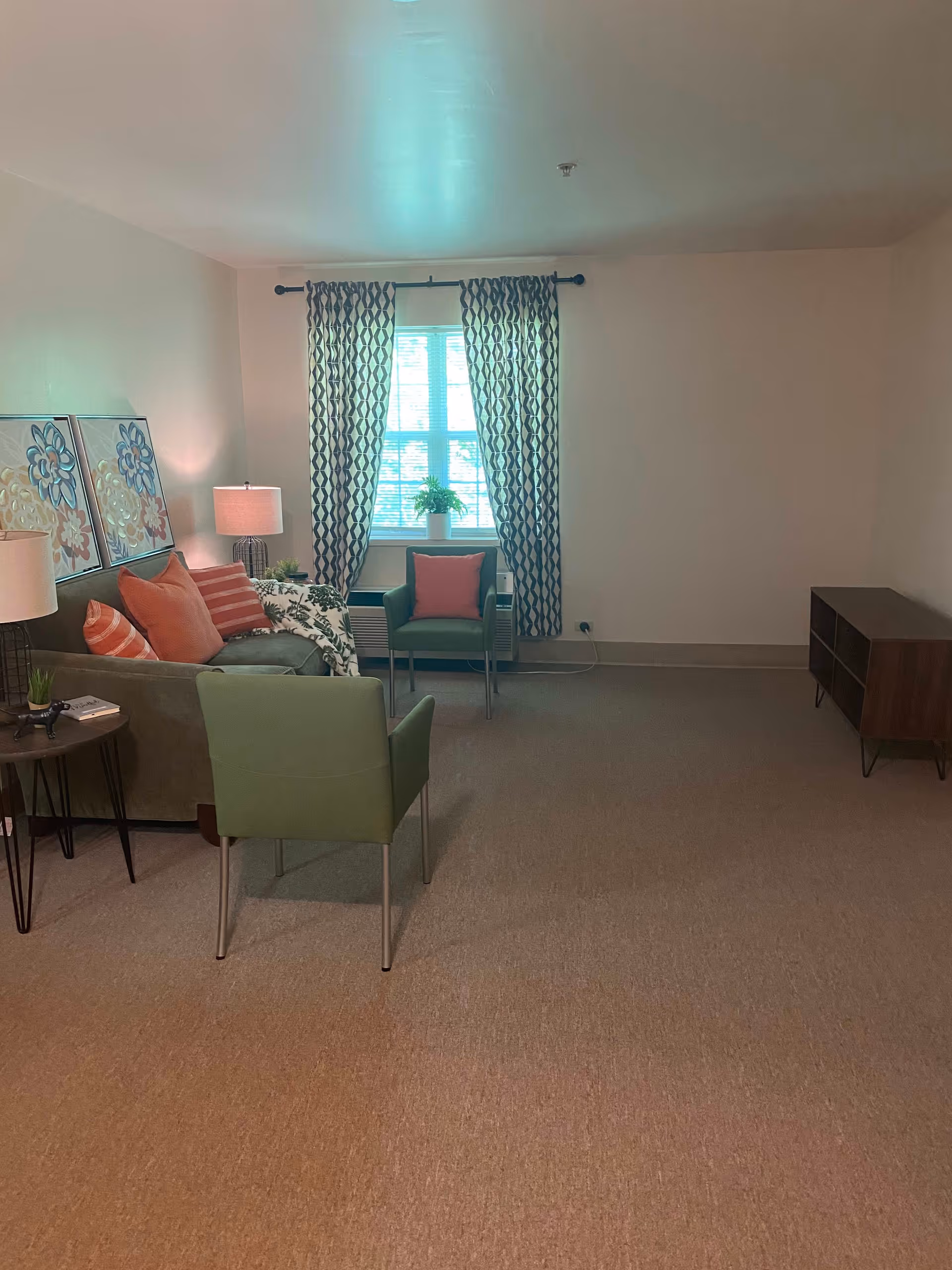 A living room with a green sofa adorned with orange and striped pillows, a green armchair with an orange pillow, a small round side table with a lamp and a plant, two floral paintings on the wall, patterned curtains covering a window, and a wooden media console against the right wall.