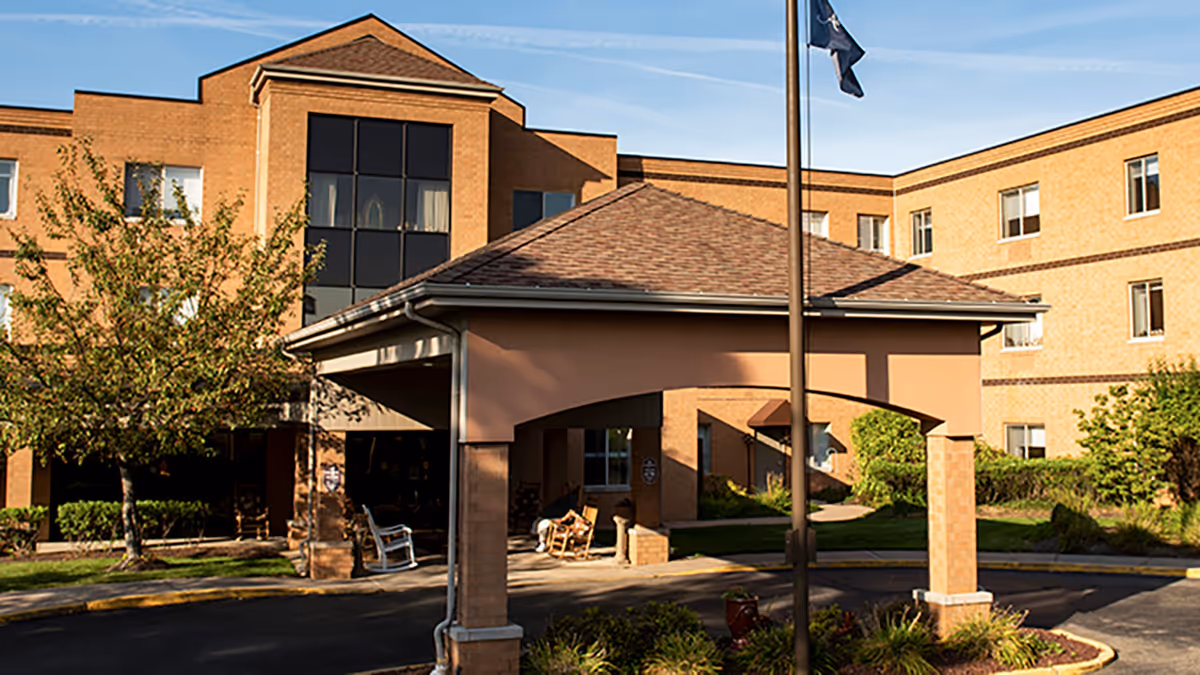 Exterior view of a multi-story brick building with a covered entrance and a flagpole in front, surrounded by trees and landscaping under a clear sky.
