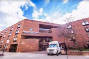 Exterior view of a multi-story brick building with a sign that reads Crown Heights Center for Nursing and Rehabilitation. A white delivery truck is parked in front of the entrance under a partly cloudy blue sky.
