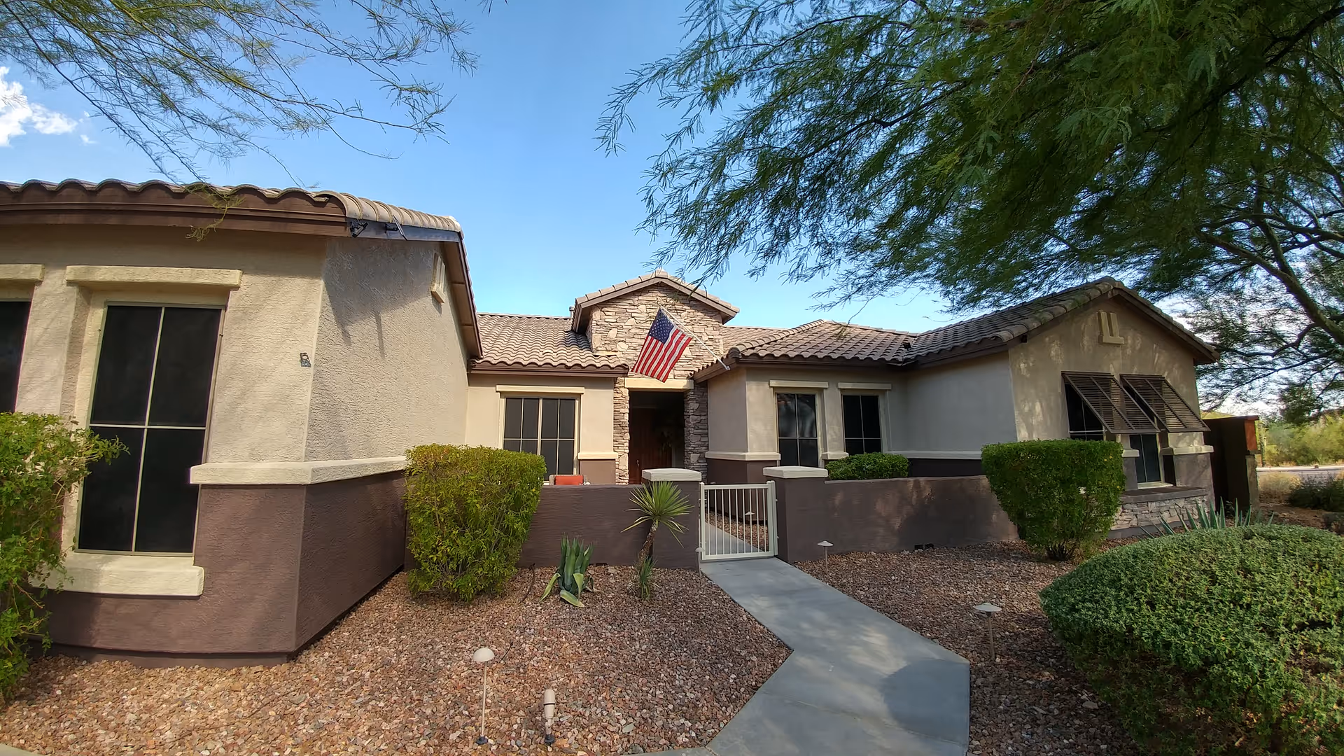 Front exterior view of a single-story senior living facility building with beige and brown walls, a tiled roof, an American flag above the entrance, and desert landscaping with bushes and gravel.