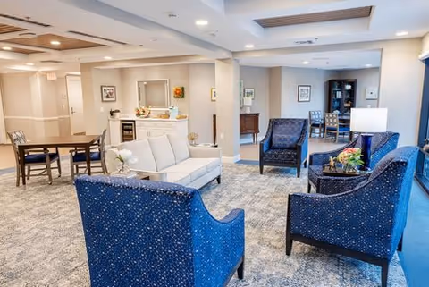 A spacious and well-lit living room area with blue upholstered armchairs and a white sofa arranged around a small table with flowers. The room features a patterned carpet, recessed lighting, and neutral-colored walls with framed artwork. In the background, there is a dining table with chairs and a small kitchenette area.