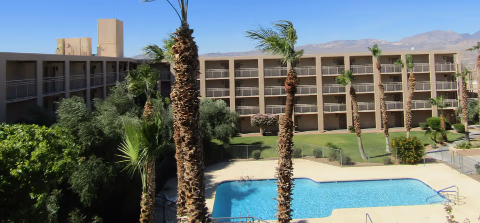 Outdoor view of a multi-story retirement facility building with balconies overlooking a swimming pool surrounded by palm trees and greenery, with mountains visible in the background under a clear blue sky.