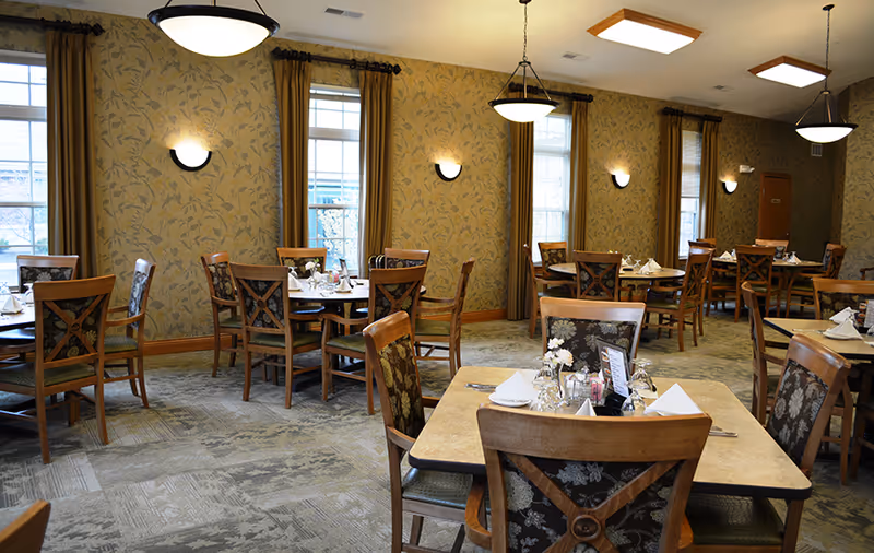 A dining room with several wooden tables and chairs arranged neatly. Each table is set with white napkins, glassware, and small flower vases. The room has patterned wallpaper, large windows with brown curtains, and ceiling lights providing warm illumination.