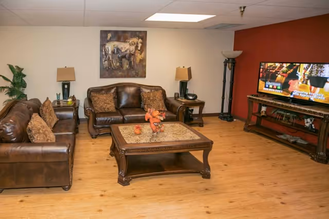 A cozy living room with two brown leather sofas, a wooden coffee table with a decorative centerpiece, two side tables with lamps, a tall floor lamp, a flat-screen TV on a wooden TV stand, and a painting of horses on the wall.