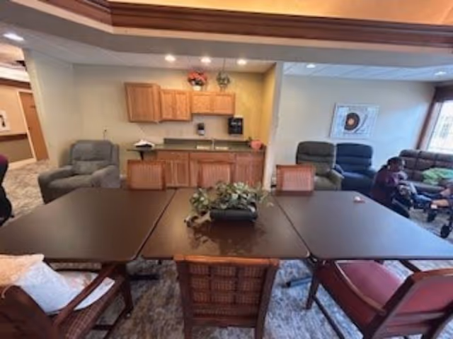 Communal dining and lounge area with a large dark table surrounded by chairs, a kitchenette and recliners in the background.