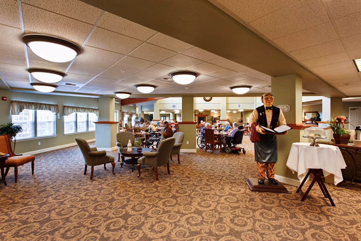 A spacious senior living community dining area with patterned carpet, several round tables with chairs, and seniors seated and dining. The room has large windows with valances, ceiling lights, and a decorative statue of a waiter holding a menu near a small table with a white tablecloth and a cake stand.