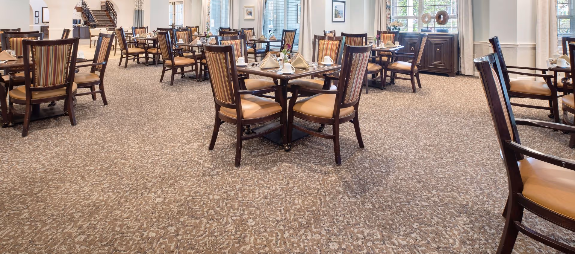 A spacious dining room with multiple wooden tables and chairs arranged neatly. Each table is set with napkins, cups, and silverware. The room has large windows with curtains, allowing natural light to brighten the space. The carpet has a patterned design, and there is a sideboard with decorative items near the windows.