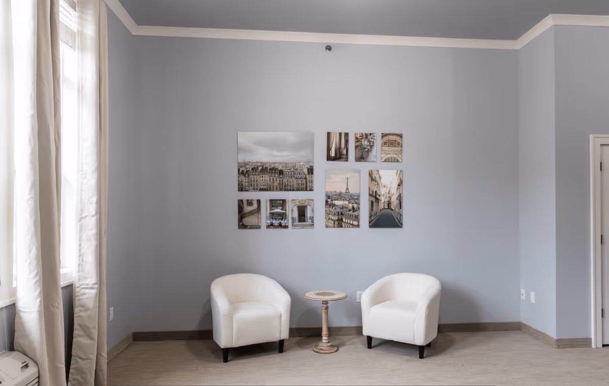 A cozy seating area with two white armchairs and a small round wooden table between them, set against a light gray wall decorated with a collage of framed photos depicting cityscapes and architectural details. Natural light filters in through a window with beige curtains on the left side.